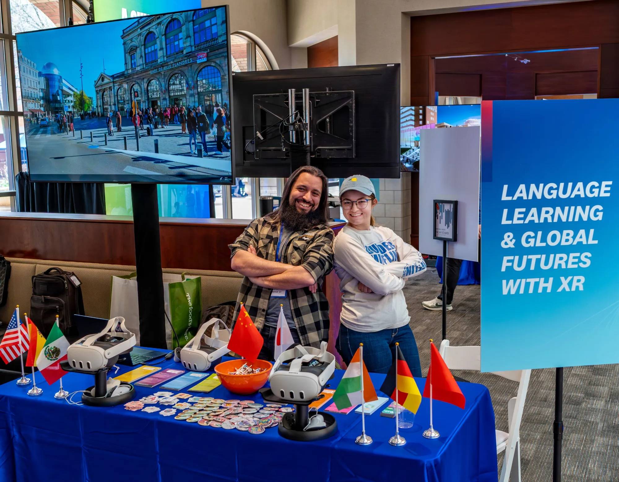 A photo from a resource fair, showing a table with international flags as well as virtual reality headsets. There is a screen in the background showing a location in europe.